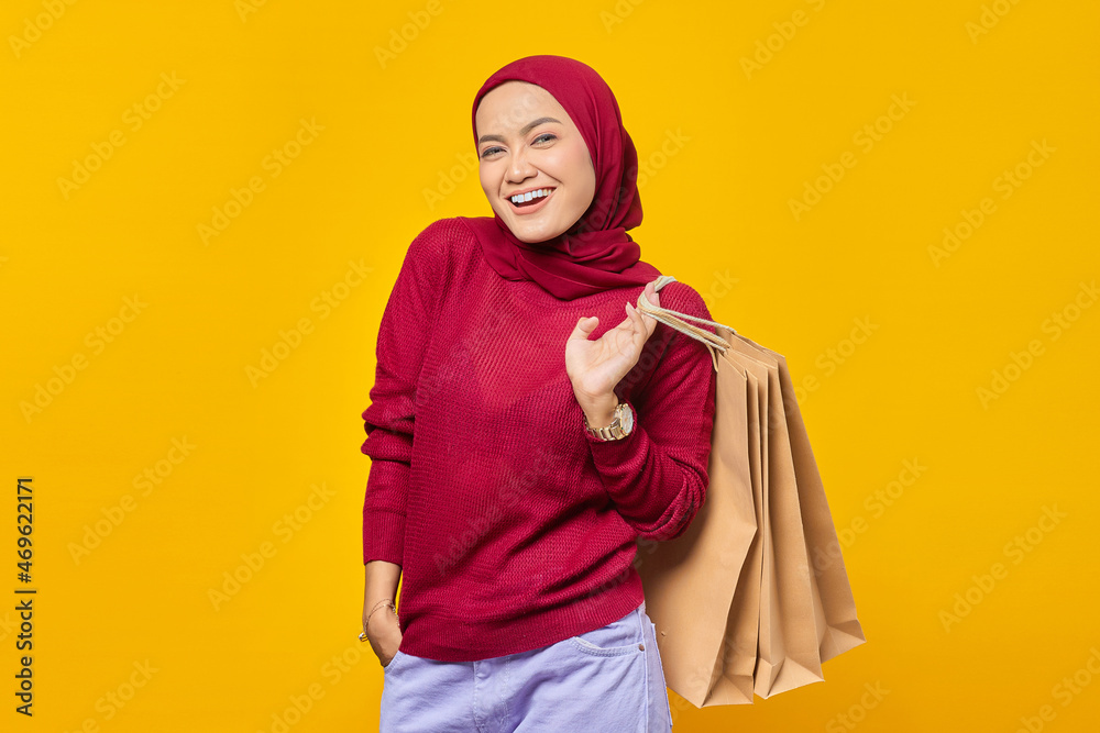 Obraz premium Happy and confident young Asian woman smiling at camera with hands in pockets and holding shopping bags on yellow background