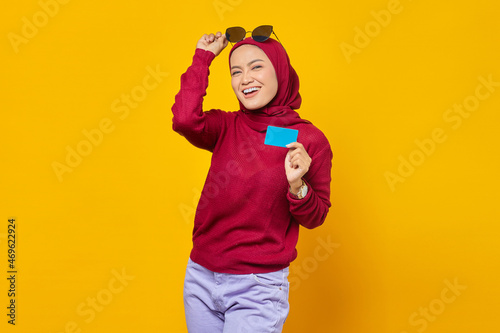 Smiling young Asian woman holding credit card and take off spectacles on yellow background