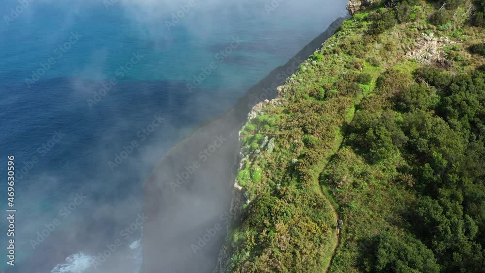 Drone shot going upwards of a young, fit and strong man hiking up Espigao Amorelo on Maderia during the summer. Thin clouds are drifting by with the blue and turquoise ocean below in the background. Drone shot going upwards of a young, fit and strong man hiking up Espigao Amorelo on Maderia during the summer. Thin clouds are drifting by with the blue and turquoise ocean below in the background.