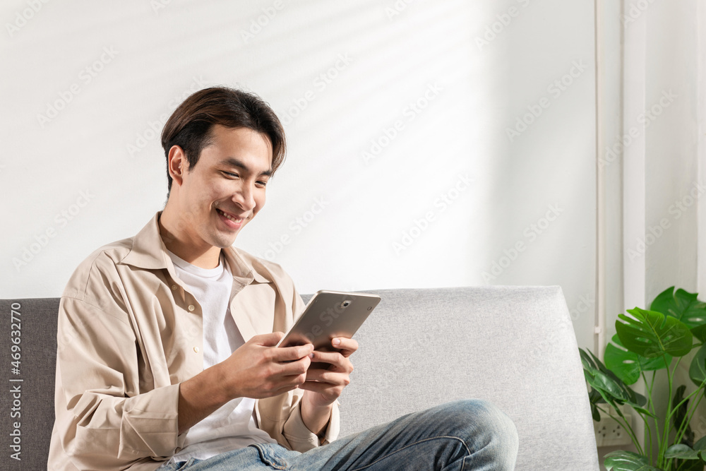 Happy smiling Asian student sits and relaxes on a couch using modern tablet browsing unlimited wireless internet, positive young man freelancer working uses wifi at a home apartment in the morning Happy smiling Asian student sits and relaxes on a couch using modern tablet browsing unlimited wireless internet, positive young man freelancer working uses wifi at a home apartment in the morning