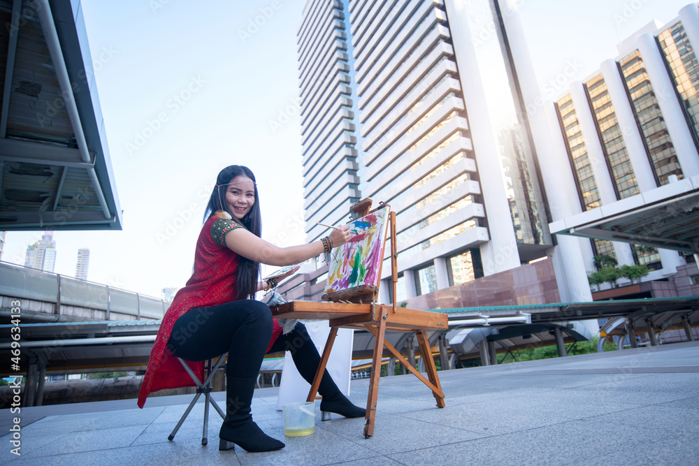 Female street artist paints a picture on the town square, modern building in background