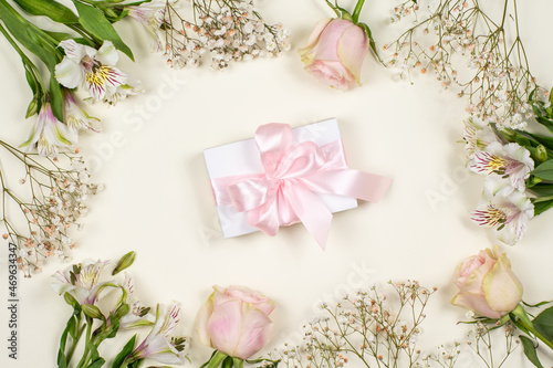 A white gift box with a pink bow on a pink background in a frame of fresh flowers. A gift for a holiday. Congratulation. A greeting card. Sale. Black Friday. Happy Birthday 