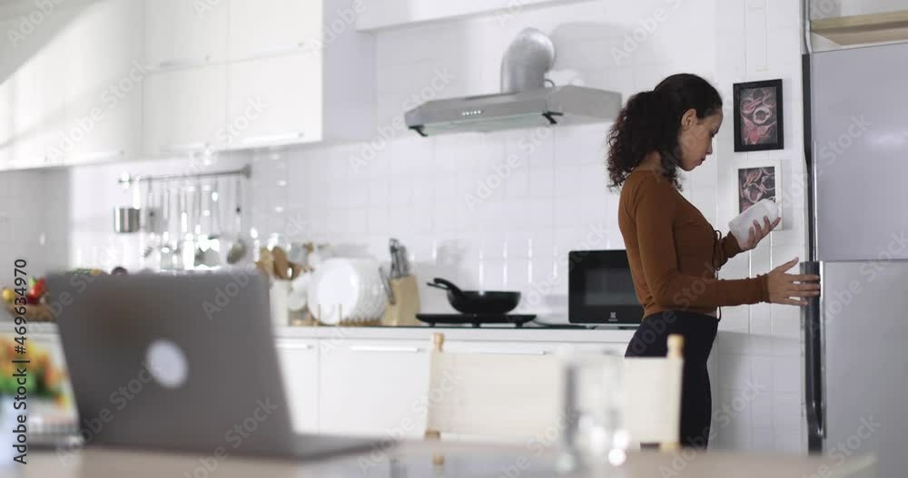 Ethnic female taking bottle of sauce from fridge and browsing website of online shop on laptop in kitchen at home Ethnic female taking bottle of sauce from fridge and browsing website of online shop on laptop in kitchen at home