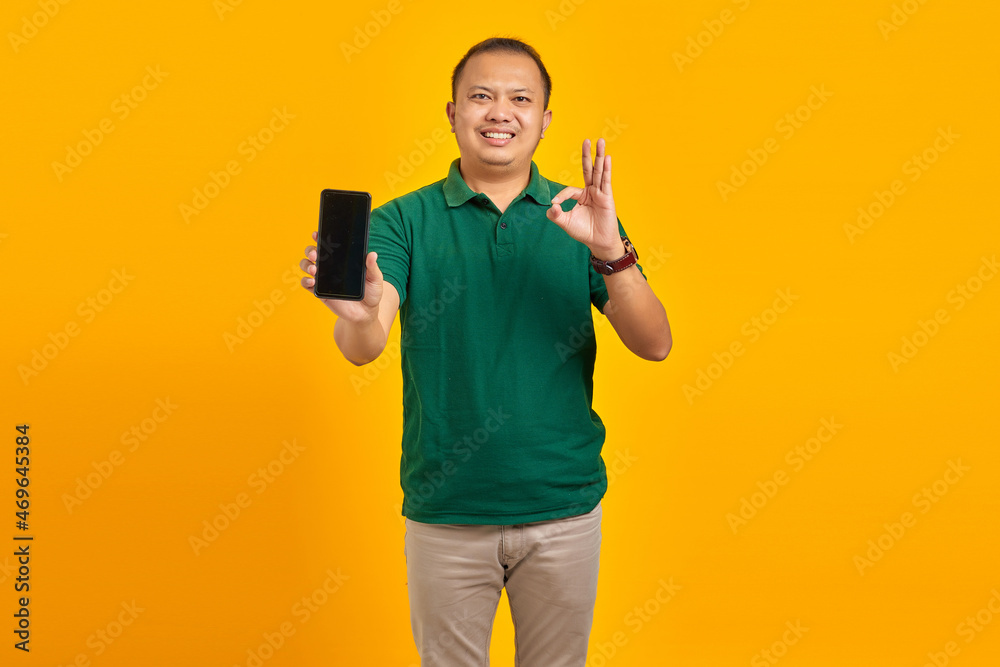 Happy Asian man showing mobile phone blank screen and hand gesture okay on yellow background Happy Asian man showing mobile phone blank screen and hand gesture okay on yellow background