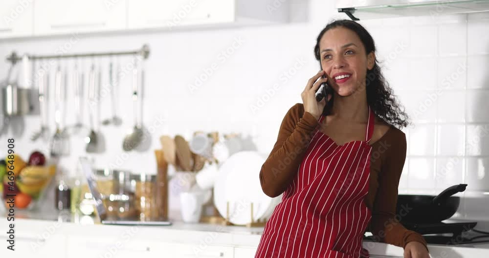 Cheerful ethnic woman in striped apron smiling and answering phone call while leaning on counter near stove in light kitchen at home Cheerful ethnic woman in striped apron smiling and answering phone call while leaning on counter near stove in light kitchen at home