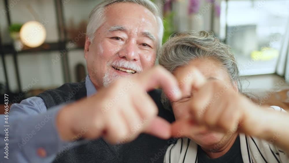 Close up portrait happy asian elderly retired family couple making heart gesture with fingers, showing love or demonstrating sincere feelings together indoors, looking at camera.