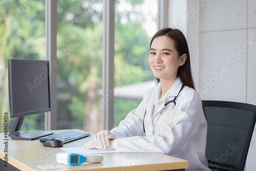 Asian professional young smiling woman doctor sitting look forward in clinic at hospital. On table has a paper and computer.
