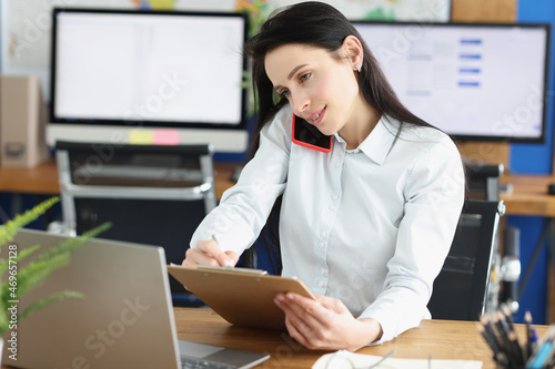 Woman worker talking on phone and making notes on paper in personal office