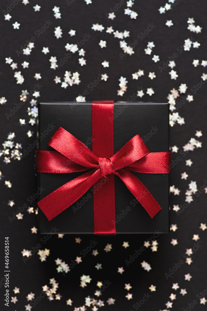 Black gift box with red ribbon on a background with silver confetti in the form of stars. Valentines, Christmas or New Year day backdrop. Flat lay style with minimalistic design.