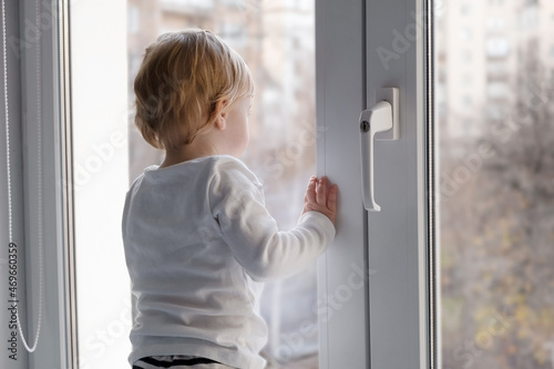 Cute caucasian blonde baby girl about 1,2 years old on sill looking through window with handle lock at street autumn view.Infant,toddler,kid safety,secuity..Lock down,isolation,stay home concept