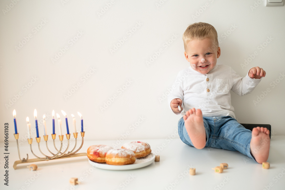 Kid in white t-shirt on white background celebrating Hanukkah. Jewish festival of lights. Child smile and happy. on table donuts and traditional menorah with lighting candles. holiday in Israel Kid in white t-shirt on white background celebrating Hanukkah. Jewish festival of lights. Child smile and happy. on table donuts and traditional menorah with lighting candles. holiday in Israel