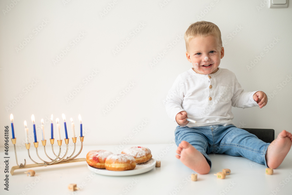 Kid in white t-shirt on white background celebrating Hanukkah. Jewish festival of lights. Child smile and happy. on table donuts and traditional menorah with lighting candles. holiday in Israel Kid in white t-shirt on white background celebrating Hanukkah. Jewish festival of lights. Child smile and happy. on table donuts and traditional menorah with lighting candles. holiday in Israel