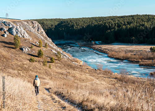 rear view of woman in neutral colors clothes walking by river in autumn on dry grass against rocks and forest, earth tones landscape hiking