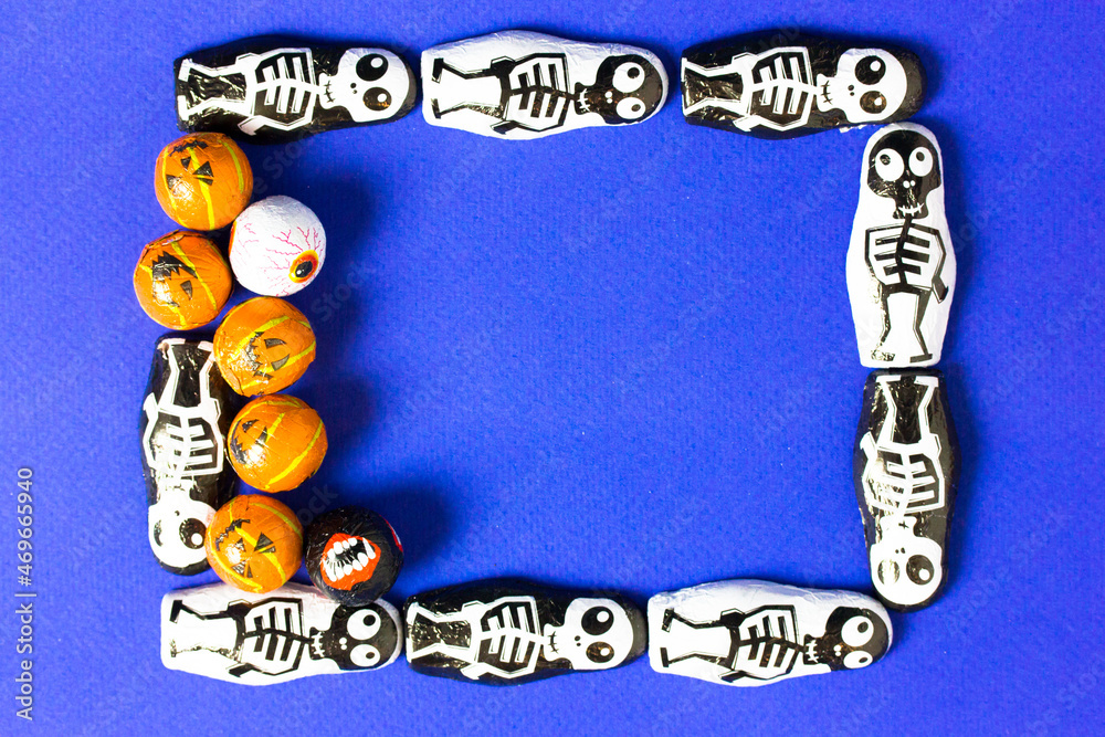 Obraz premium Creative frame of black and white skeleton candies and orange jack o lantern pumpkins with space for text on blue background. Halloween celebration concept. Invitation to a party on October 31st