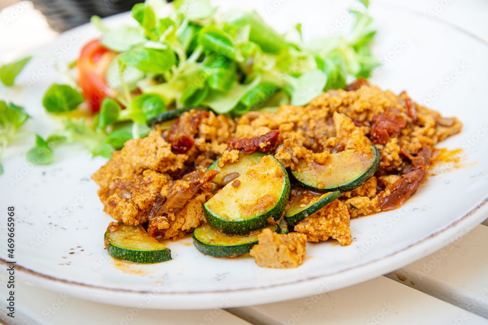 Vegan breakfast prepared with scrambled tofu and eggs, cooked with dried tomatoes and zucchini served with fresh lettuce and cucumber in white ceramic vintage plate