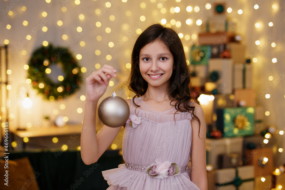 Fototapeta premium cute girl in festive dress holding christmas decoration in decorated room with heap of christmas gifts