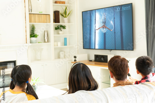 Rear view of family sitting at home together watching swimming competition on tv
