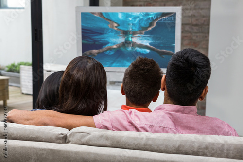 Rear view of family sitting at home together watching swimming competition on tv