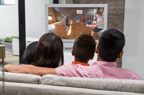Rear view of family sitting at home together watching basketball match on tv