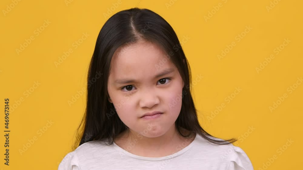 Little grumpy asian girl pouting lips, grimacing and looking angrily to camera, close up portrait, orange background Little grumpy asian girl pouting lips, grimacing and looking angrily to camera, close up portrait, orange background