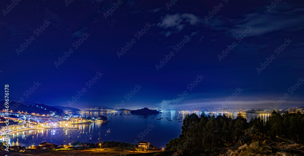 Panorámica de pueblos costeros de noche en la Ría de Pontevedra con playa, embarcadero, barcos,  bateas de mejillones y isla y ciudad de fondo con luces.
