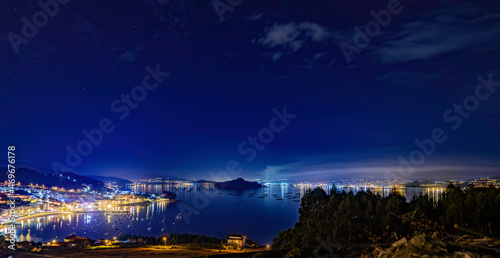 Panorámica de pueblos costeros de noche en la Ría de Pontevedra con playa, embarcadero, barcos,  bateas de mejillones y isla y ciudad de fondo con luces.