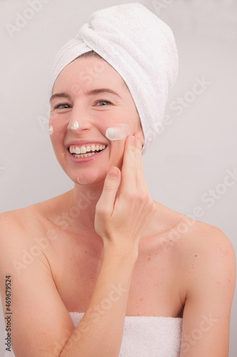 Close up beauty portrait of a laughing woman applying facial cream with a towel on her head.