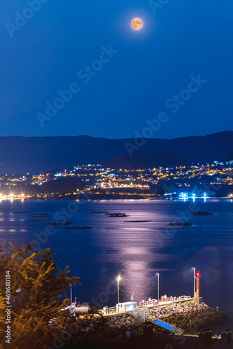 Embarcadero en la ría de Pontevedra de noche con la luna y ciudad de fondo con luces y bateas de mejillones