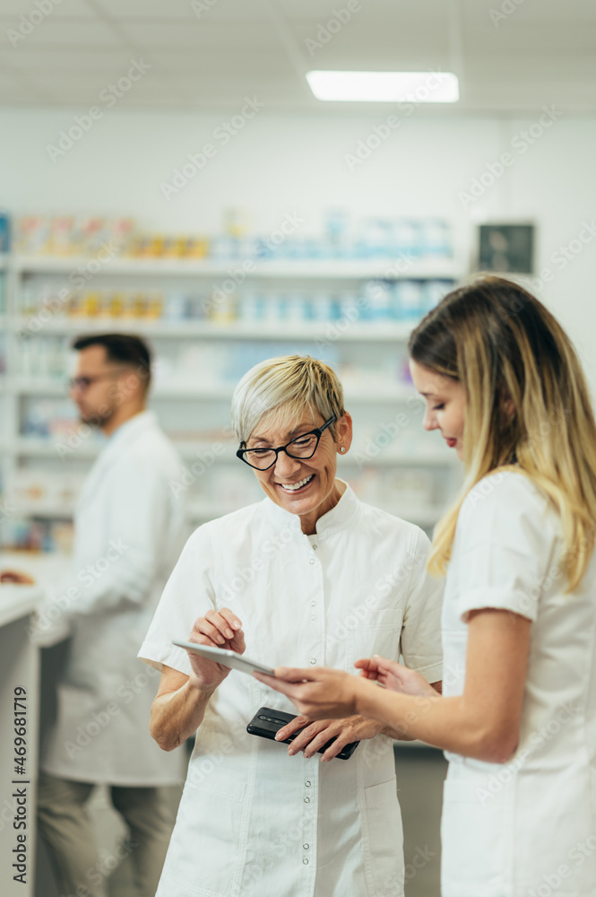 Obraz premium Senior female pharmacist using a tablet while working in a pharmacy with her young colleague