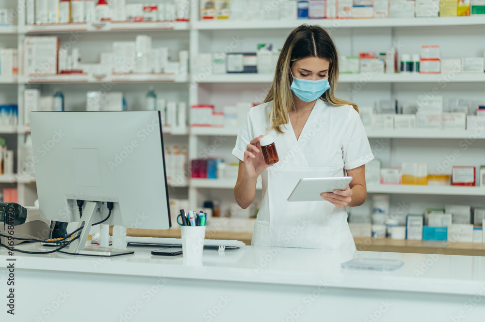 Pharmacist with protective mask on her face holding drugs and using digital tablet