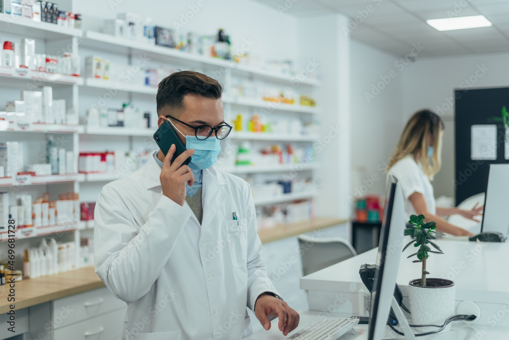 Pharmacist with protective mask on his face using a smartphone while working at a pharmacy