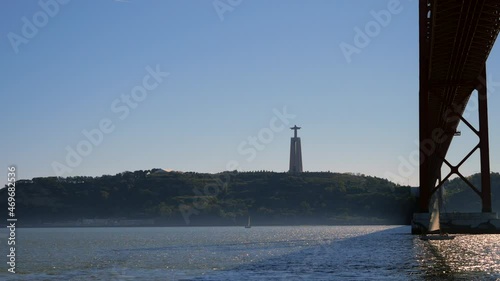 A yacht passing under the 25 de Abril Bridge and overlooked by the Cristo Rei statue in Lisbon, Portugal. Filmed in 4K