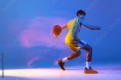 Dynamic portrait of professional basketball player training with ball isolated on blue studio background in mixed neon light.