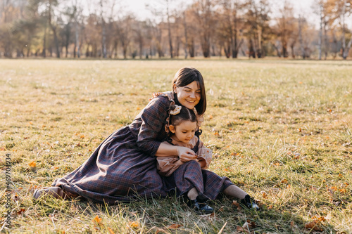 Mother and daughter spending time outdoors in a park. Woman and little girl sitting on green grass in a field, wearing matching outfits.