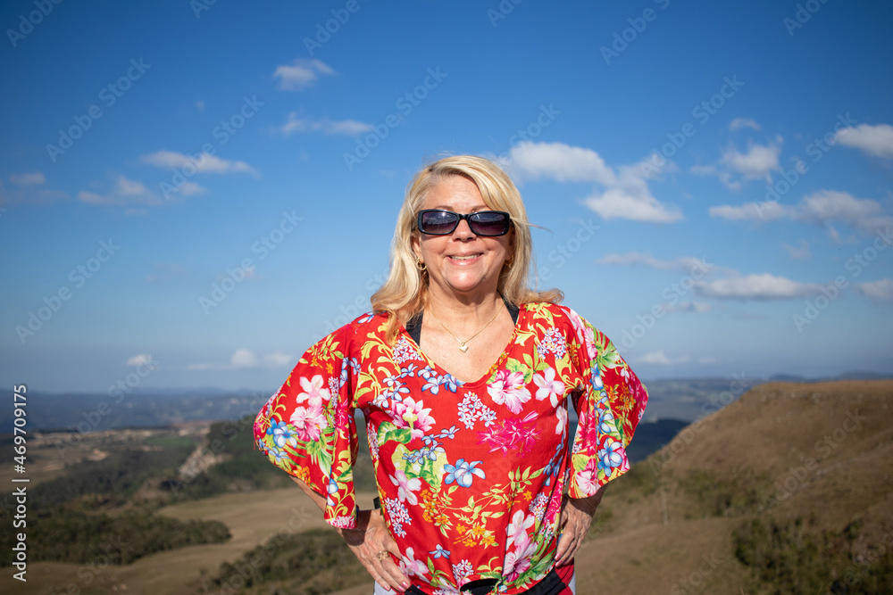 blonde lady with printed blouse and sunglasses, looking at photo and smiling, in the background beautiful landscape, space for text