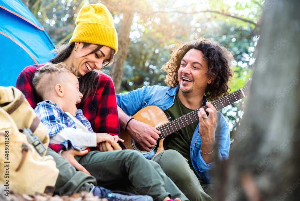 Happy family camping in the forest playing guitar and singing together - Mother, father and son having fun trekking in the nature sitting in front of the tent - Family, nature and trekking concept. Happy family camping in the forest playing guitar and singing together - Mother, father and son having fun trekking in the nature sitting in front of the tent - Family, nature and trekking concept.