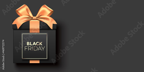 Black Friday gift vector banner, with a premium style black gift box wrapped with golden satin ribbon, and lettering label, lying on background with copy space.