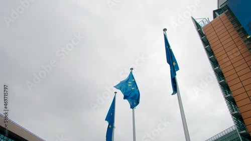 Establishing shot of three waving EU Flags on gray sky background near European Commission Headquarters in Brussels, Belgium. Political, Financial or Economics Crisis Concept. 4K medium wide shot photo