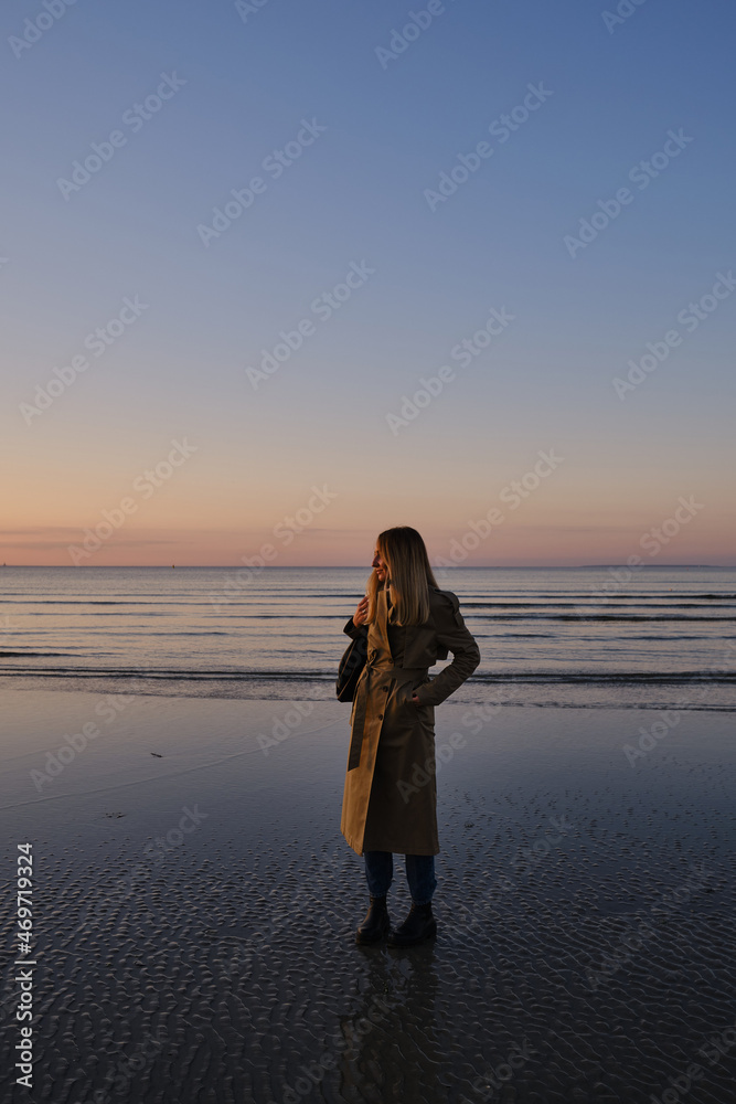 Fototapeta premium Stylish girl walks at sunset along the beach of the sea or ocean on a cool day. Travel concept. Walk on the sea or ocean. Girl in travel. Amazing scenic outdoors view. Beautiful sunset on the seashore