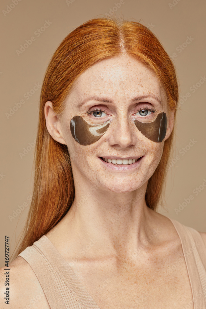 Vertical portrait of beautiful red haired woman with freckles smiling at camera while enjoying skincare using moisturizing patches Vertical portrait of beautiful red haired woman with freckles smiling at camera while enjoying skincare using moisturizing patches