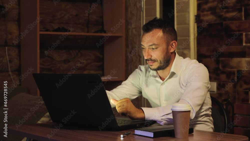 Handsome businessman in white smiles, he made a profit. A young man with a beard works at home using a laptop. Business concept remotely, online. 4K UHD. Handsome businessman in white smiles, he made a profit. A young man with a beard works at home using a laptop. Business concept remotely, online. 4K UHD.