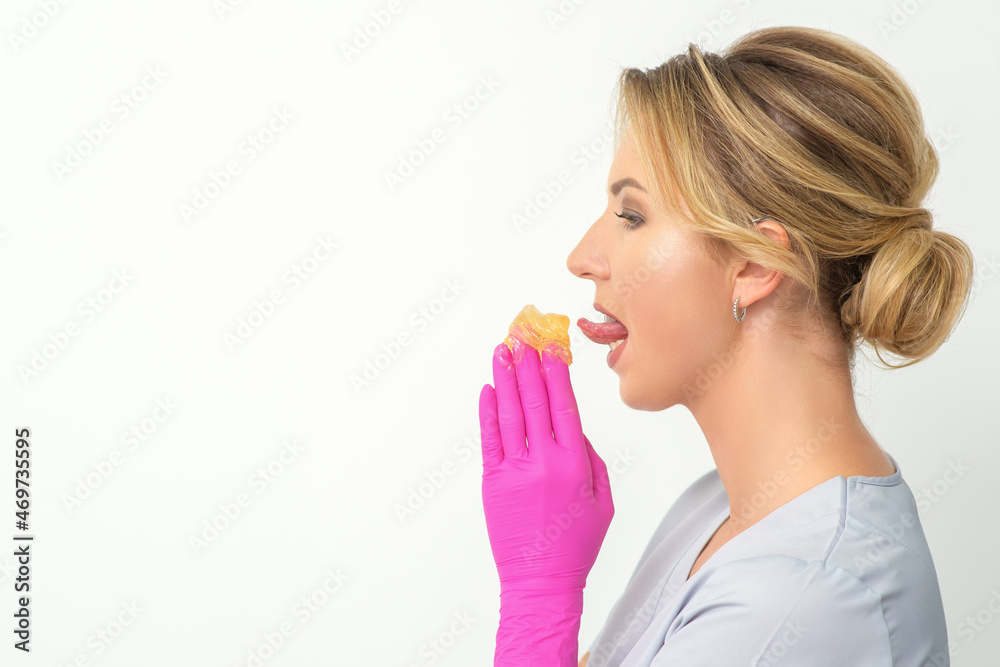 Young beautician licks liquid wax for depilation holding in gloved hands standing on white background. Cosmetologist tastes the sugar paste to taste Young beautician licks liquid wax for depilation holding in gloved hands standing on white background. Cosmetologist tastes the sugar paste to taste