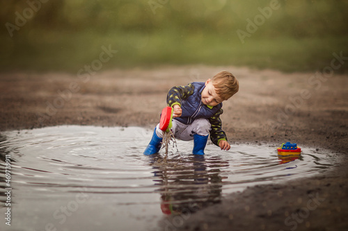 Boy in rain boots crouching in puddle and playing with ships photo