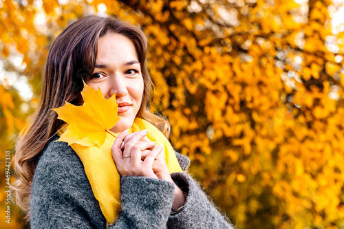 Portrait of a young woman holding a maple leaf in front of her and smiling in the park on a walk. She is dressed in a gray polto and a yellow scarf. Autumn discount and sale. Space for text