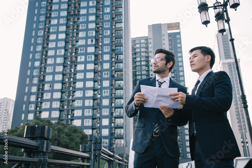 Businessman leadership use smartphone and talking with assistant while hold paper sheets of project outside near cityscape and dreaming for bigger projects with business assistants outside office. 