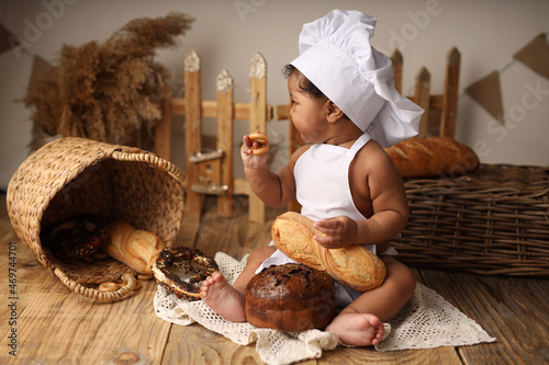 a cute dark-skinned kid with curly hair in a chef's costume has a bagel and a roll. High-quality photography