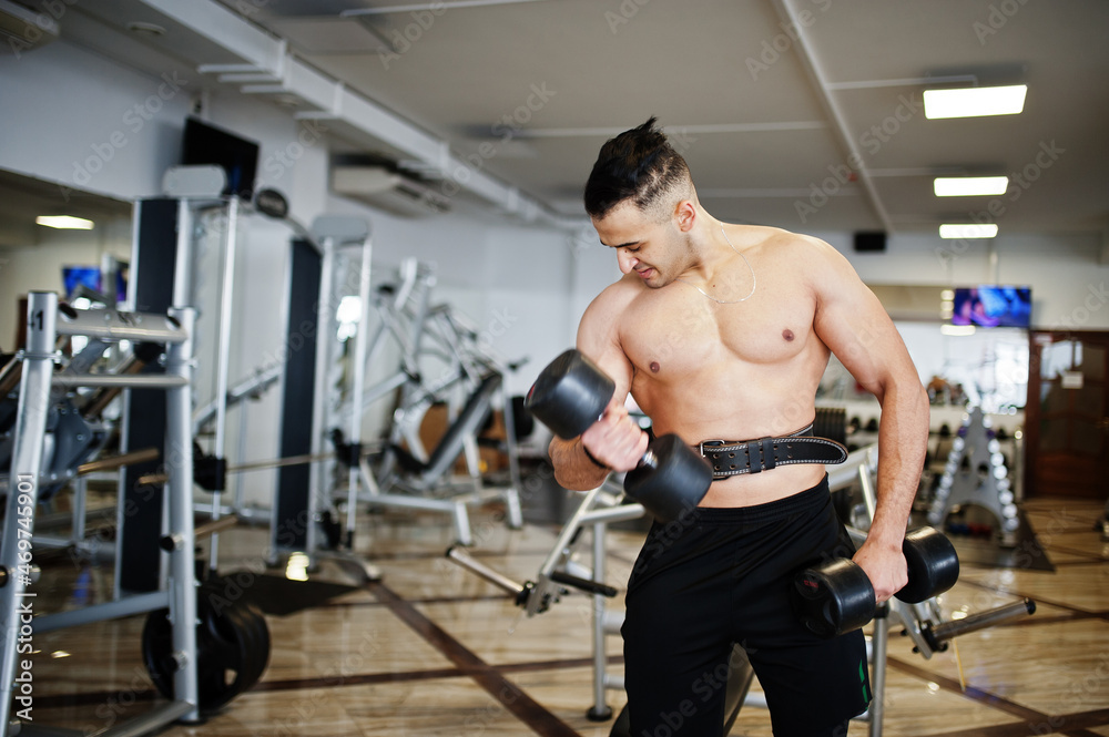 Fototapeta premium Muscular arab man training in with dumbbells modern gym. Fitness arabian men with naked torso doing workout .
