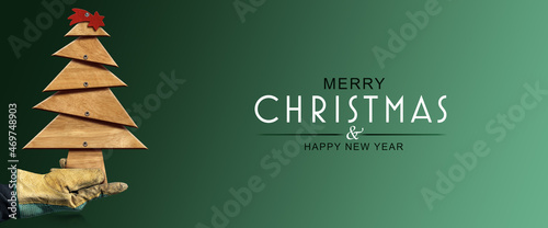 Hand with protective work gloves holding a small wooden Christmas tree with a red comet star, on green background with text Merry Christmas & Happy New Year.