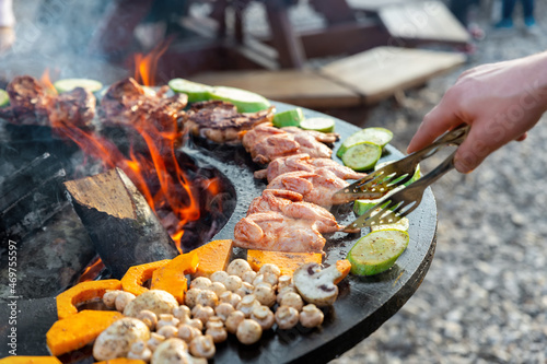 Close-up detail view of chef flipping by tongs tasty crispy marinated quails and vegetables grilled at round steel iron firepit hearth table surface brazier with burning firewoods. Barbecue yard home photo