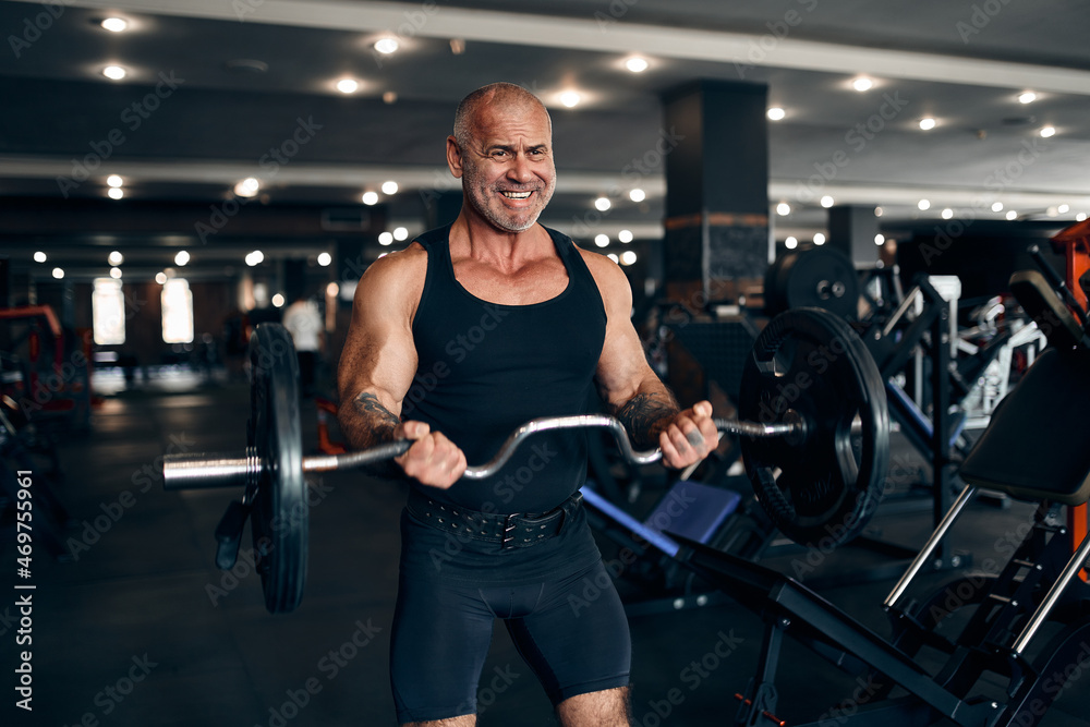 Muscular older man working out in gym doing exercises with barbell at biceps. Bodybuilding concept Muscular older man working out in gym doing exercises with barbell at biceps. Bodybuilding concept